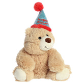 A soft brown bear plush toy from Aurora with a festive birthday hat and a sweet smile.