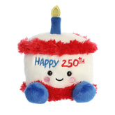 A small red, white, and blue cake plush with “Happy 250th” embroidery and a gold candle from Palm Pals by Aurora.