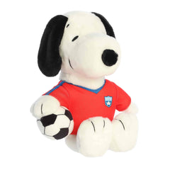 A Snoopy plush wearing a red soccer jersey and holding a soft soccer ball from the Peanuts collection by Aurora.