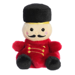 Adorable, mini nutcracker plush toy sitting with their red uniform, curly mustache, and tall hat