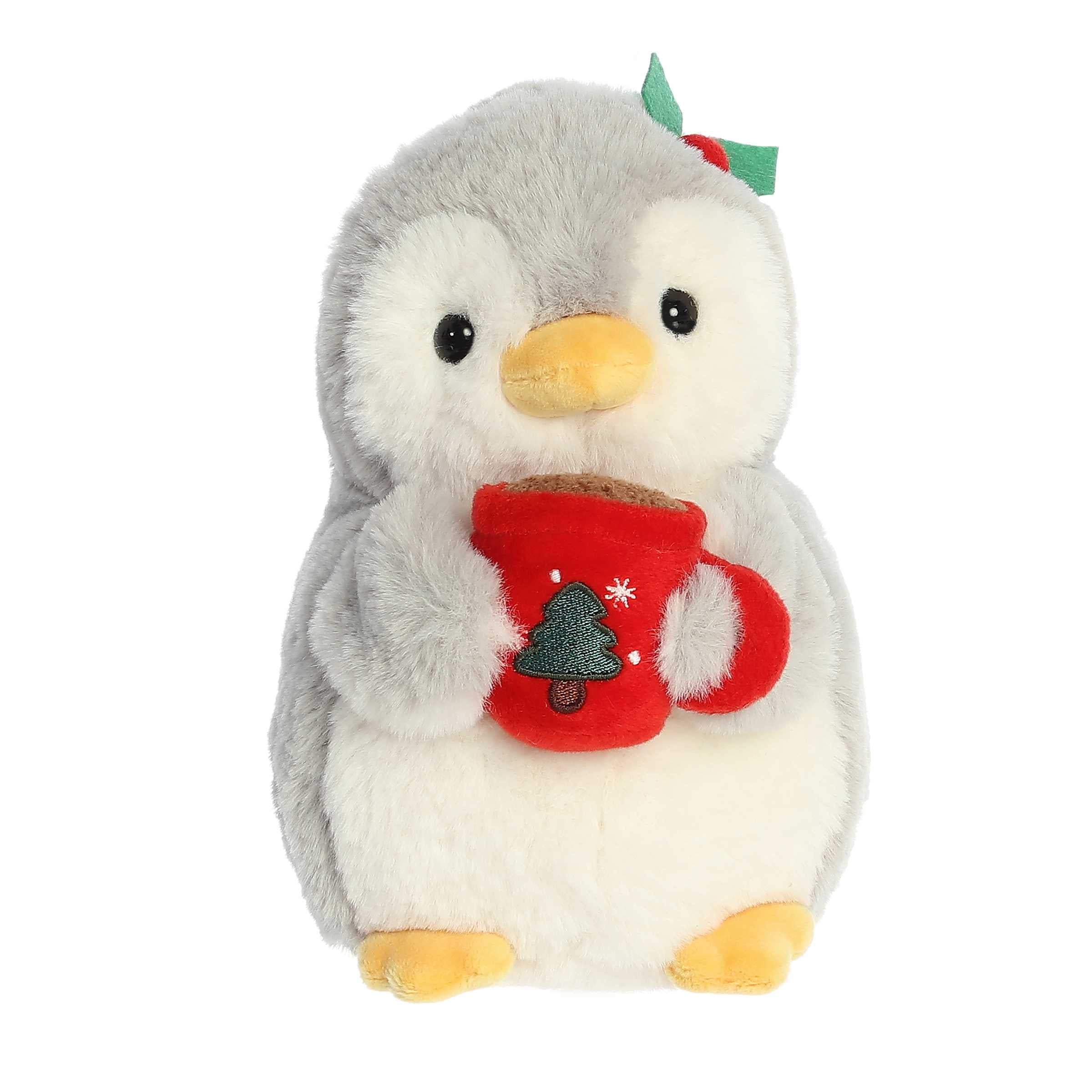 Light grey penguin with a yellow beak and feet wearing holly on it head holding a red mug with a Christmas tree in front